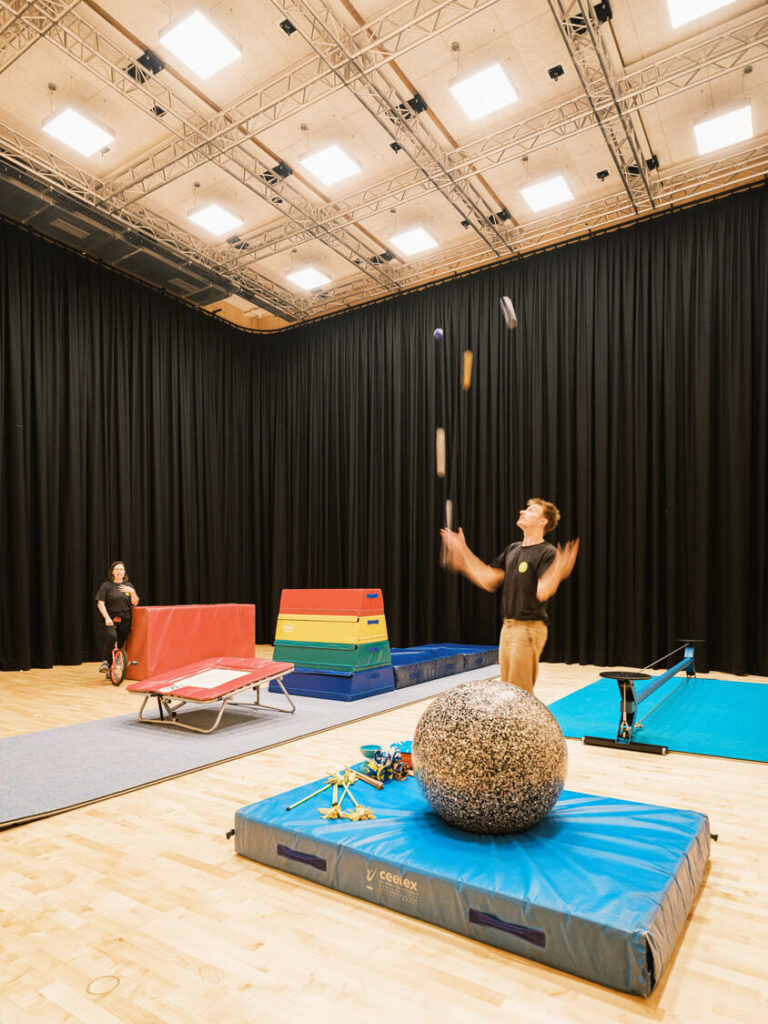 A man juggles against a black curtain, surrounded by acrobatic equipment.