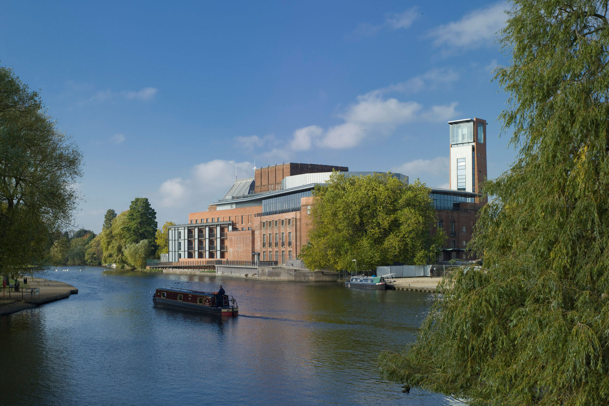 A boat cruises up the river behind the brick Royal Shakespeare Theatre building.