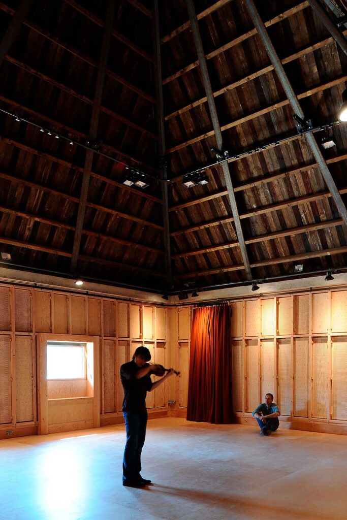 A person plays a violin while another looks on under the tall, angled roof.