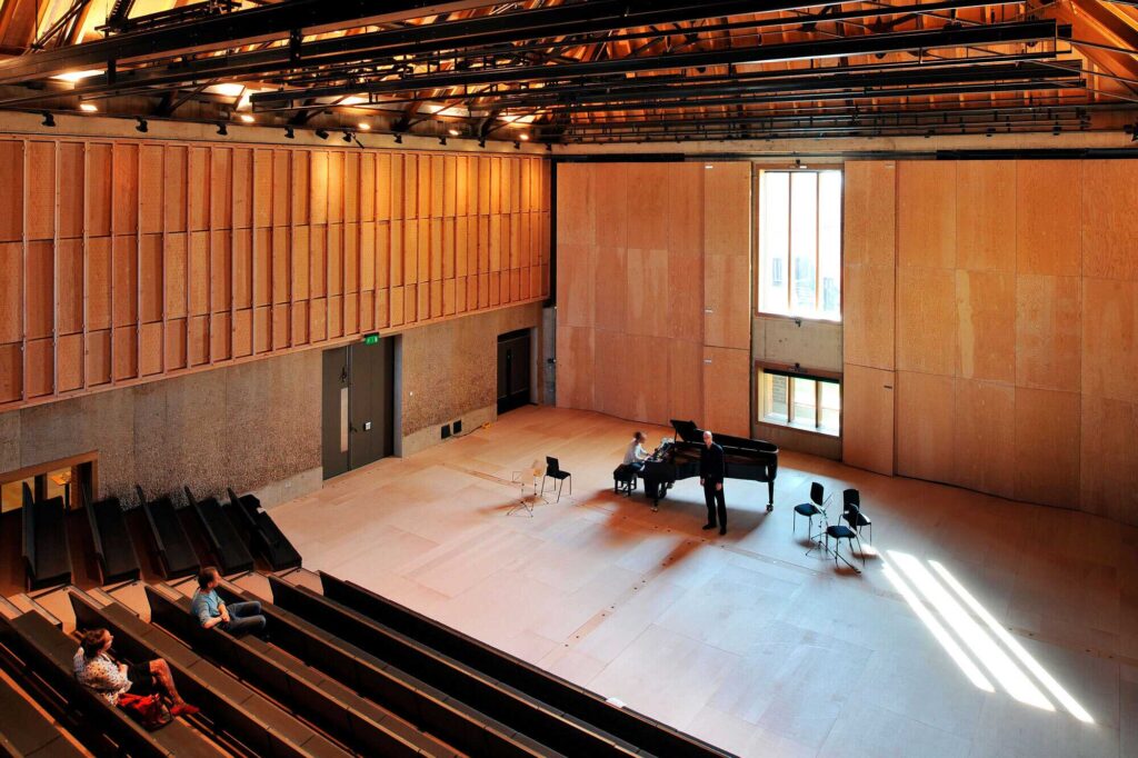 People practice around a piano in a large rectangular performance space.