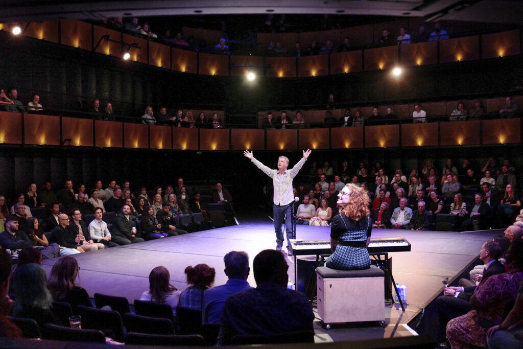 A man raises hands on stage while a woman looks up from a keyboard. Image by Haworth Tompkins.