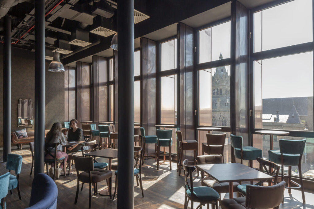 Two women sit in a cafe or lounge area with tall windows overlooking a street.