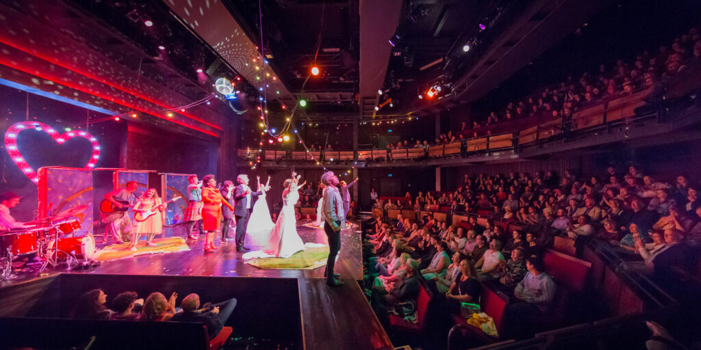 Numerous performers face the audience. The stage is strung with colorful lights and a band plays guitar and drums.