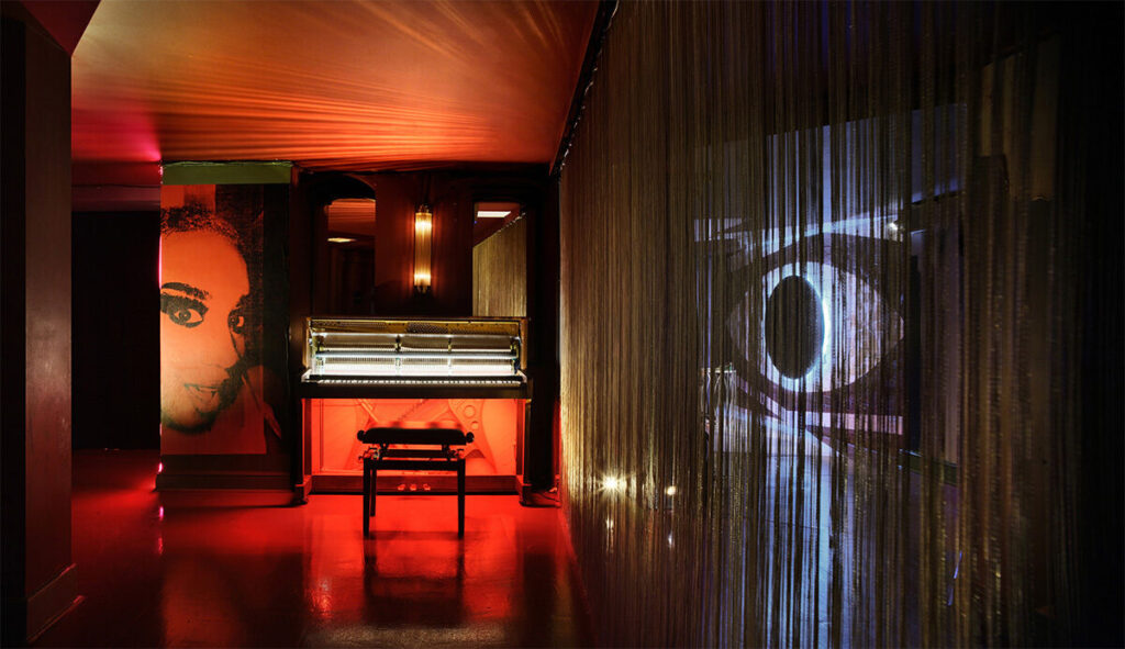 A curtain of threads divides a piano from a room with an eye shaped mirror.