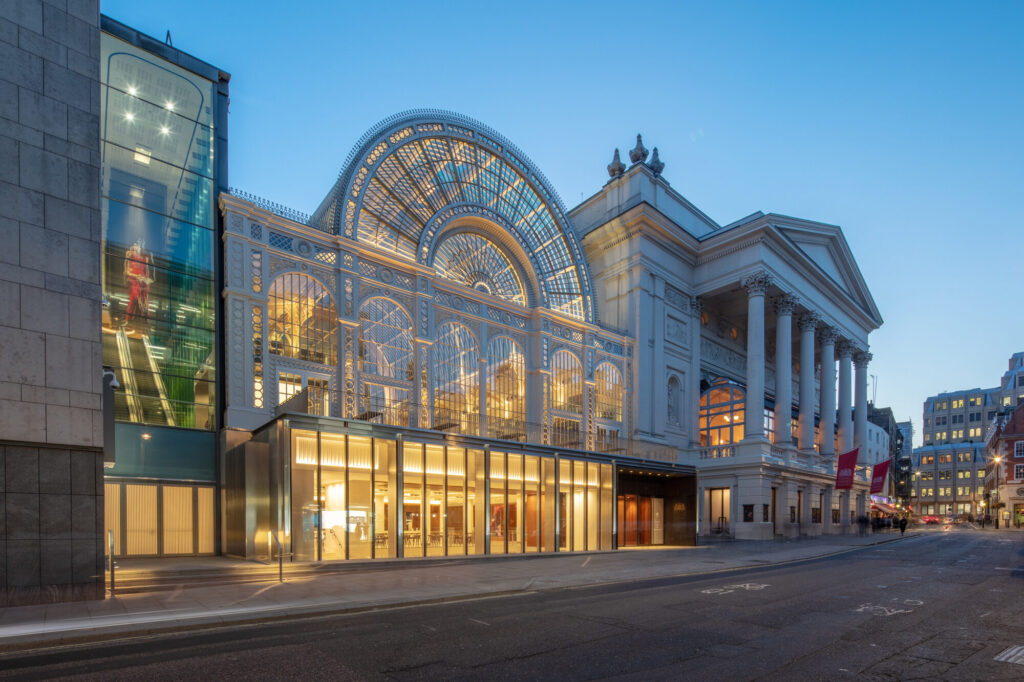 Glass exterior of The Linbury Theatre Royal Opera House illuminated gold against a blue evening sky.
