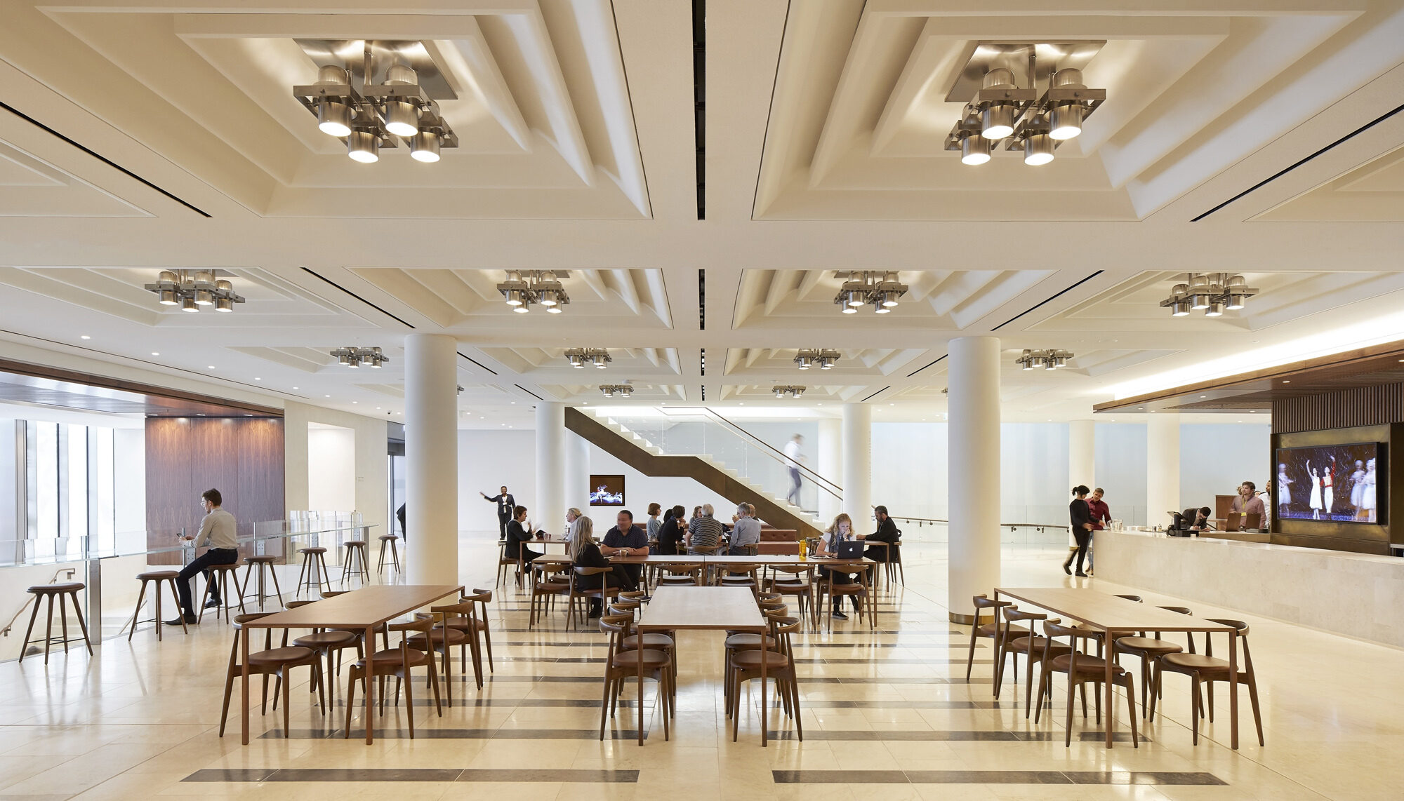 A white and bronze cafeteria-style seating area with stairwell in the background and bar or cafe area on the right.