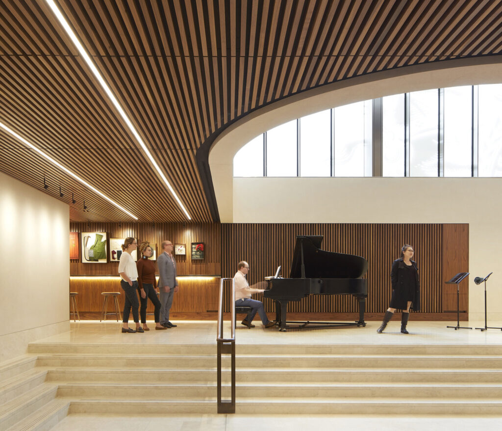 A man plays piano on a stairwell landing. Windows let in natural light above and a seating area continues off to the left.
