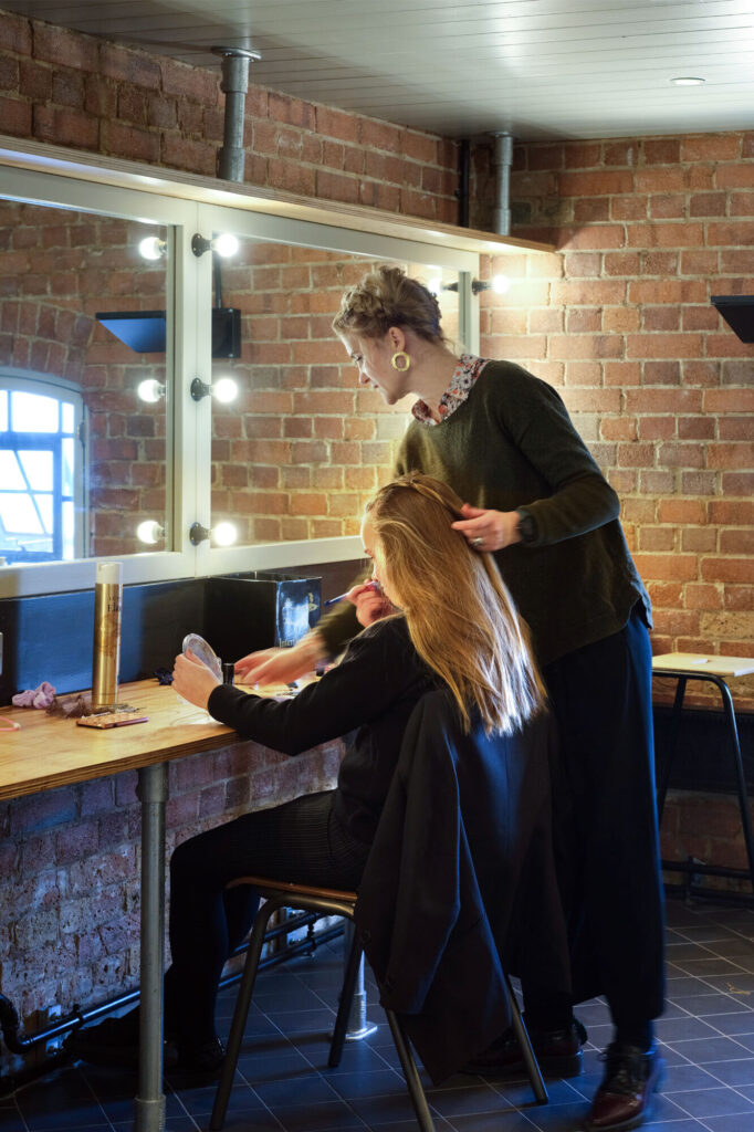 A girl looks in a mirror as an a woman braids her hair.