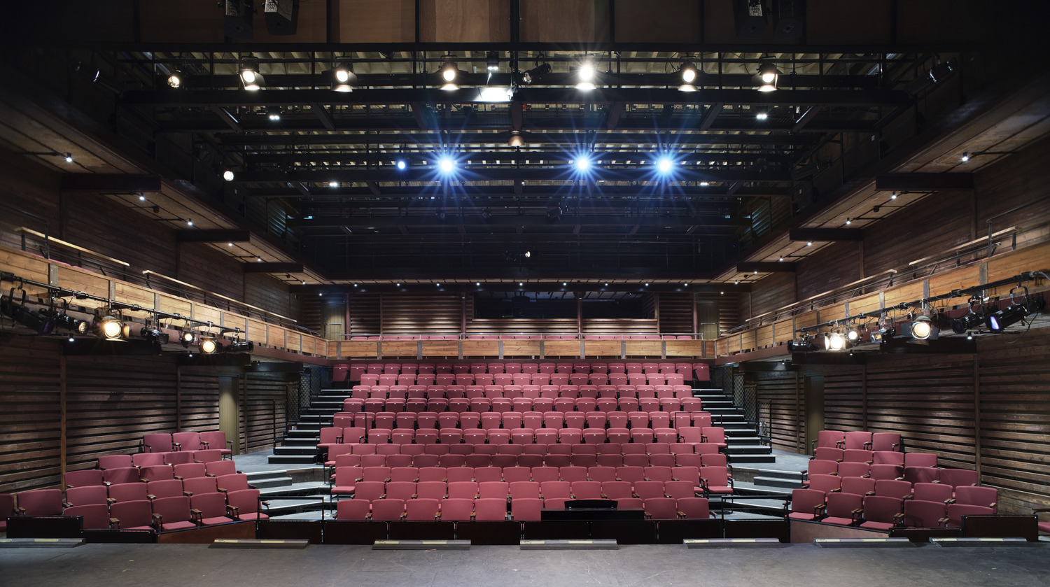 Two levels of seating with stairs and balcony level. The red seats are framed with rustic wood wall paneling.