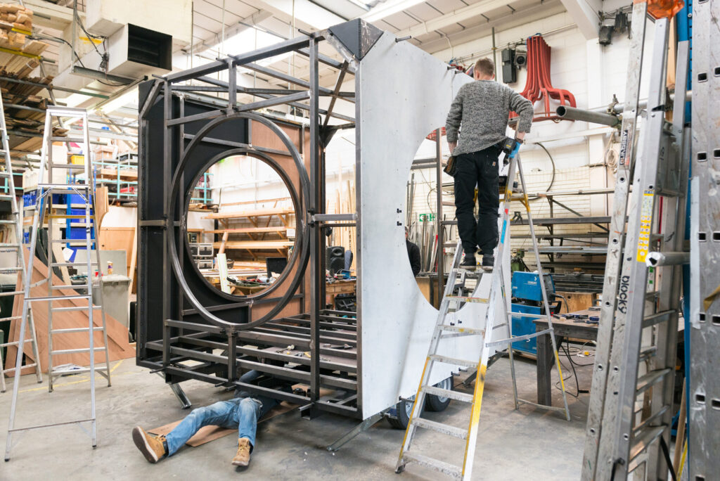In a workshop a man stands on a ladder screwing a panel onto the metal frame of the Pod. Another person lays on the floor, working on the bottom of the Pod.