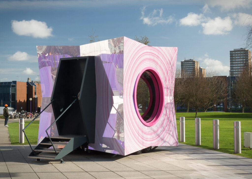 Reflective, geometric cube shaped trailer, with foldable stairs extended. It sits in a plaza surrounded by bollards and grass.