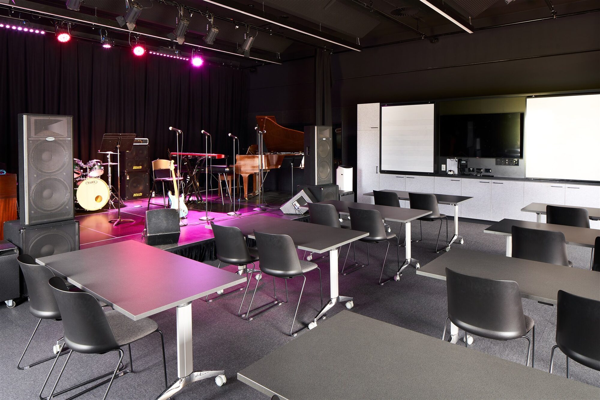 A classroom setup with tables and chairs. On the left a stage is configured with drums, guitar, keys, a piano and large speakers.