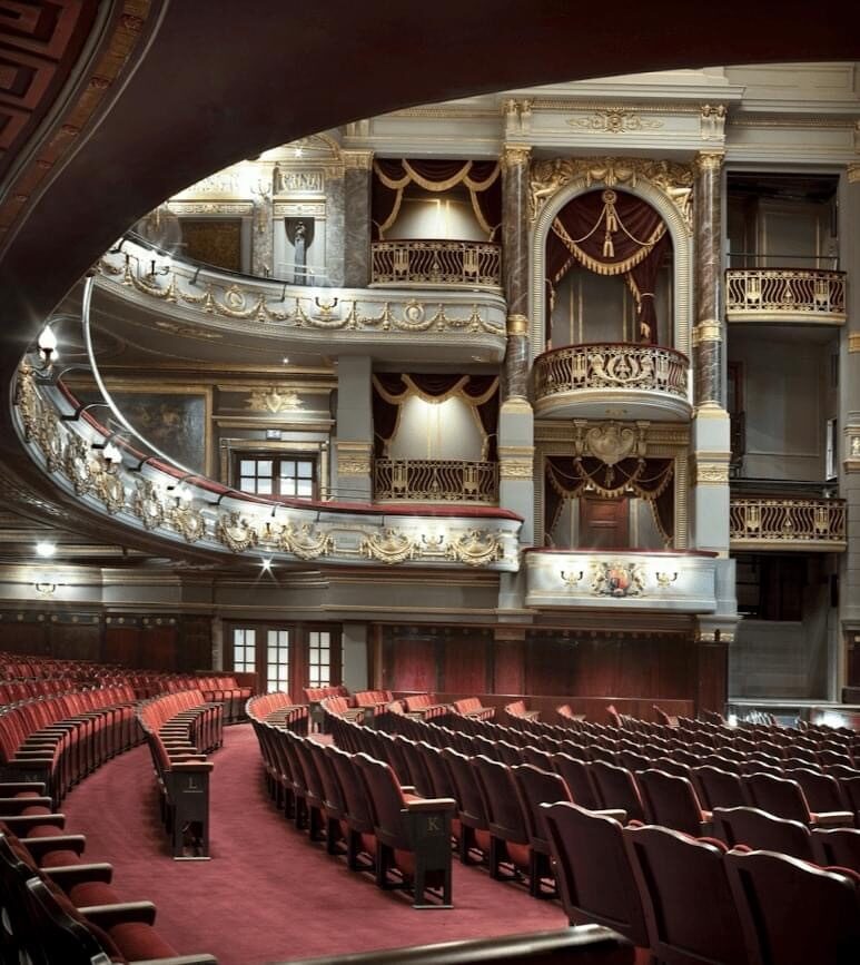 View from the red-carpeted orchestra seating area looking up toward heavily decorated white and gold balcony levels.