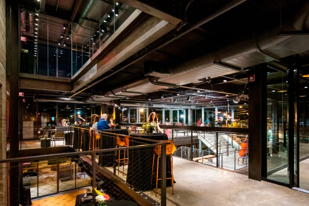 People lounge on a balcony level in the TheatreSquared lobby at night.