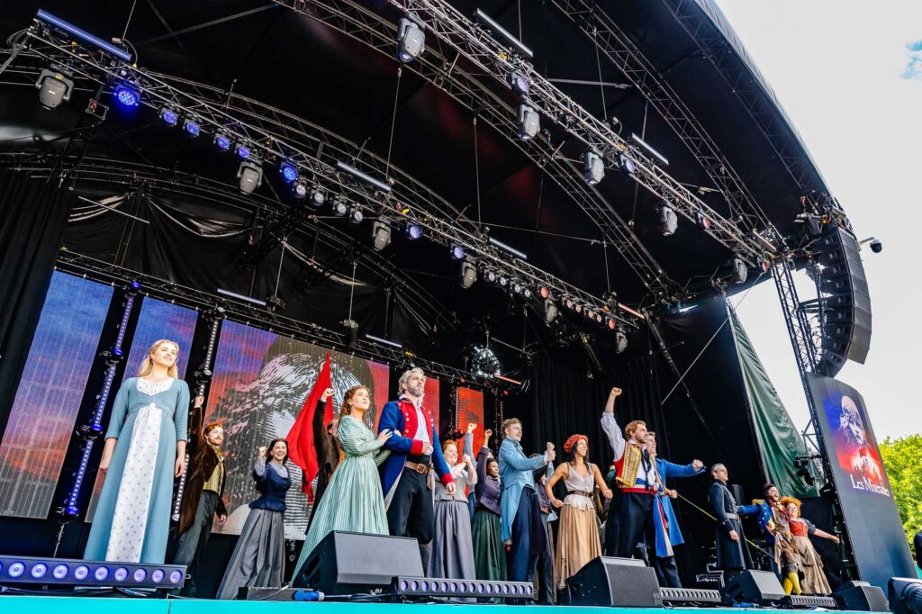 Les Miserables actors in costume pose as a group with arms raised.
