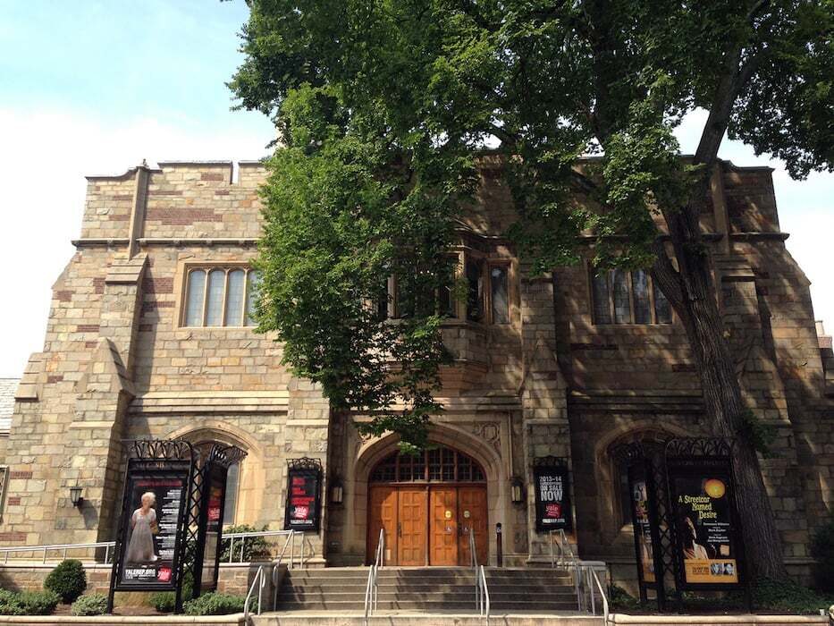 Historical stone building with theatre promotional signage flanking the entrance and a tree in front.