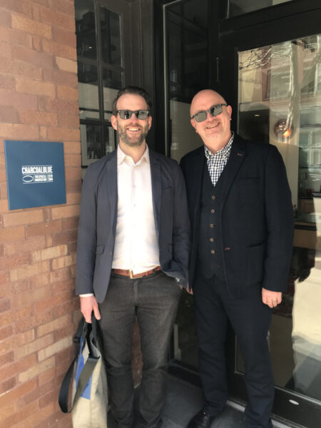 Two men in sunglasses stand outside by a door.